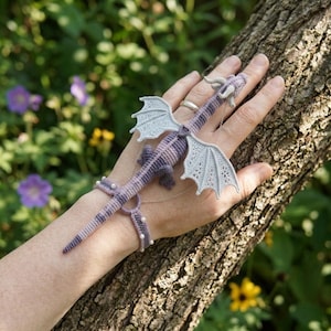 May include: A crochet dragon hand ornament featuring a purple and grey striped body with intricate light grey lace wings. The dragon is connected by thin silver chains to a matching crochet wrist cuff adorned with small white beads.