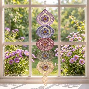 May include: A vertical hanging mobile featuring five interconnected, almond-shaped frames in a gradient of pastel colors, each containing a crocheted purple flower with a yellow center. The structure is suspended by a string from the top.