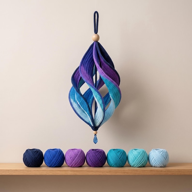 May include: A hanging spiral ornament crafted from crocheted yarn in a gradient of blue and purple shades, featuring a wooden bead at the top and a blue crystal drop at the bottom, displayed above seven matching yarn balls in a row.