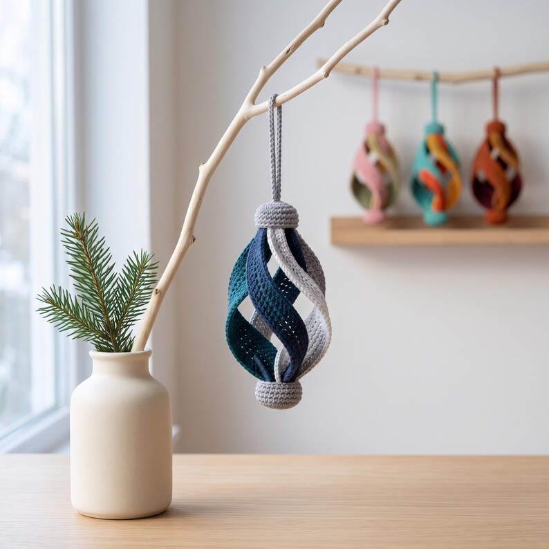 Pode incluir: Um ornamento de croch&ecirc; suspenso com um design de gaiola em espiral em tons de azul-petr&oacute;leo, azul-marinho e cinza claro. O ornamento possui uma tampa de croch&ecirc; cinza s&oacute;lida no topo e na base, com uma al&ccedil;a cinza correspondente para pendurar.