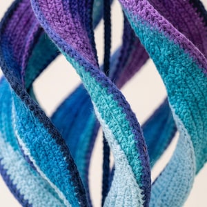 May include: A spiraling, three-dimensional sculpture crafted from crocheted yarn. The structure features a gradient of colors including deep purple, royal blue, turquoise, and light sky blue, arranged in vertical, twisted, and interconnected bands.