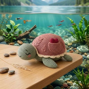 Könnte beinhalten: Ein gehäkeltes Schildkrötenspielzeug mit salbeigrünem Kopf und Gliedmaßen, einer altrosa Schale mit strukturiertem Blattmuster, einem kleinen roten Herzdetail und einem cremefarbenen, gewellten Rand. Die Schildkröte hat gestickte Augen.