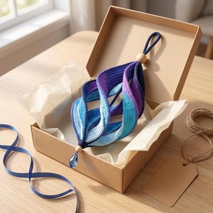 May include: A spiral-shaped crochet ornament featuring a gradient of dark blue, purple, and light blue yarn. The piece includes a wooden bead at the top and bottom, with a faceted blue crystal drop hanging from the base, all inside a cardboard box.
