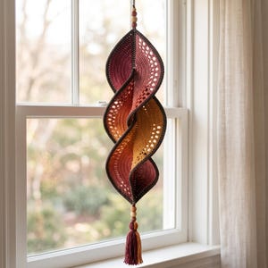 May include: A hanging spiral crochet decoration featuring a gradient of colors transitioning from deep burgundy to warm orange and yellow. The piece is constructed with intricate openwork stitches, finished with wooden beads at both ends and a tassel.