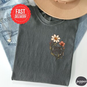 May include: A charcoal grey t-shirt featuring a floral graphic on the chest with the text "GROW GENTLY" in a dark script font. The design includes a large white daisy, a smaller orange flower, and green leaves, printed on a short-sleeve crew neck shirt.