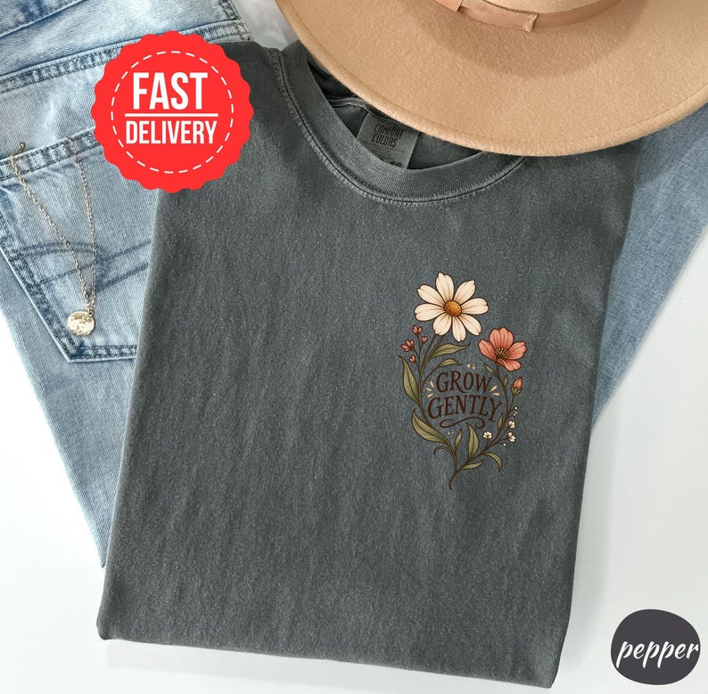 May include: A charcoal grey t-shirt featuring a floral graphic on the chest with the text "GROW GENTLY" in a dark script font. The design includes a large white daisy, a smaller orange flower, and green leaves, printed on a short-sleeve crew neck shirt.