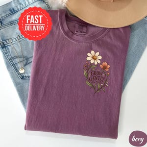May include: A plum-coloured t-shirt featuring a graphic of two flowers with green leaves and stems on the left chest. The text "GROW GENTLY" is printed in a dark script font between the flowers. The shirt has a crew neckline and short sleeves.