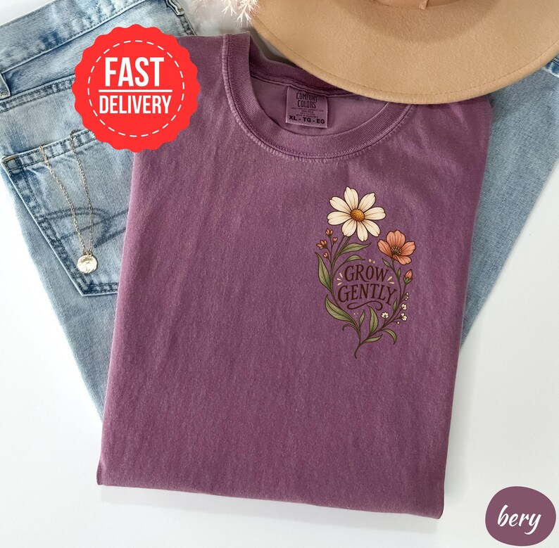 May include: A plum-coloured t-shirt featuring a graphic of two flowers with green leaves and stems on the left chest. The text "GROW GENTLY" is printed in a dark script font between the flowers. The shirt has a crew neckline and short sleeves.