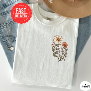 May include: A white t-shirt featuring a floral graphic on the chest with the text "GROW GENTLY" in a dark brown script font. The design includes a large cream-coloured daisy and a smaller coral-coloured flower with green leaves and stems.
