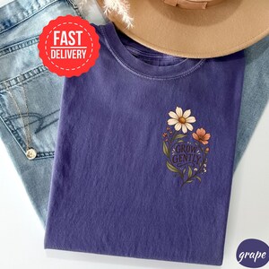 May include: A grape-coloured t-shirt featuring a floral graphic on the chest with the text "GROW GENTLY" in a script font. The design includes a large white daisy and a smaller orange flower with green leaves and stems on a dark purple fabric.