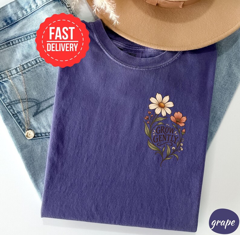May include: A grape-coloured t-shirt featuring a floral graphic on the chest with the text "GROW GENTLY" in a script font. The design includes a large white daisy and a smaller orange flower with green leaves and stems on a dark purple fabric.