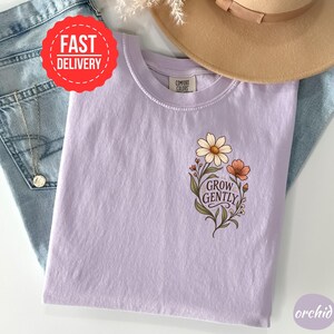 May include: A light purple t-shirt featuring a floral graphic on the chest with the text "GROW GENTLY" in a dark script font. The shirt has a crew neckline and a Comfort Colors brand label. The design includes a large white daisy and a smaller orange flower with green leaves and stems.