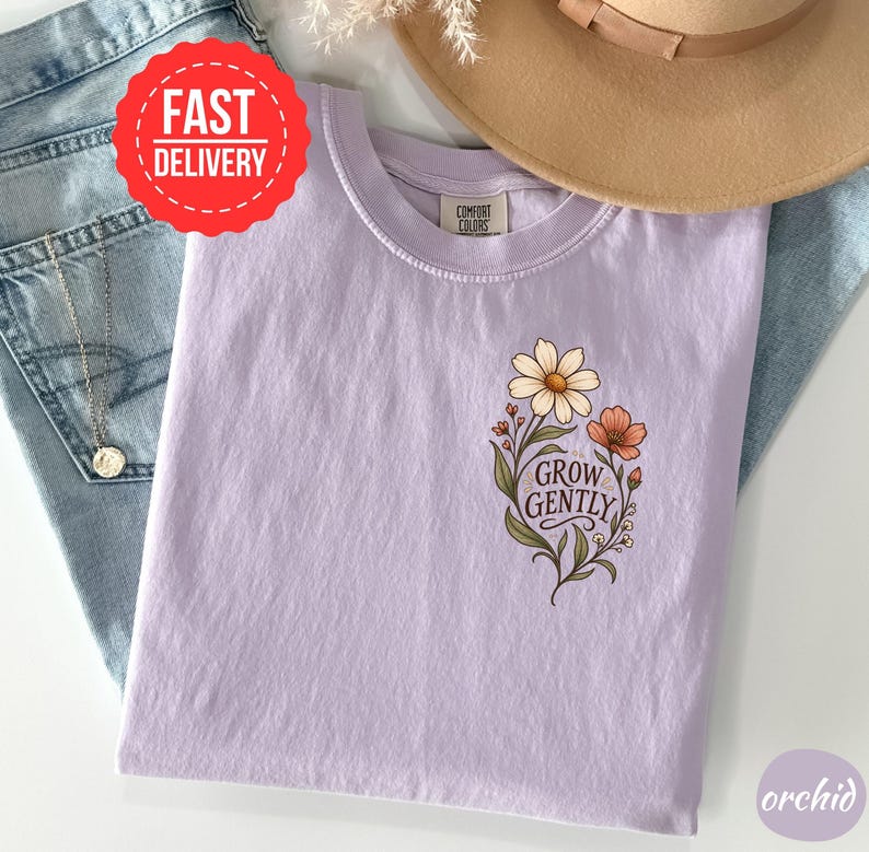 May include: A light purple t-shirt featuring a floral graphic on the chest with the text "GROW GENTLY" in a dark script font. The shirt has a crew neckline and a Comfort Colors brand label. The design includes a large white daisy and a smaller orange flower with green leaves and stems.