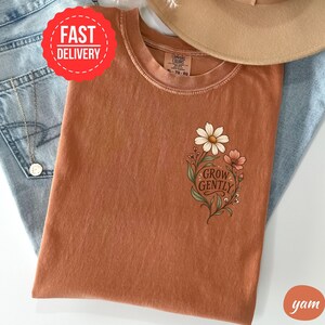 May include: A rust-coloured crew neck t-shirt featuring a floral graphic on the chest with the text "GROW GENTLY" in a dark brown script font. The shirt has a Comfort Colors label at the inner neckline and is displayed alongside blue denim fabric.