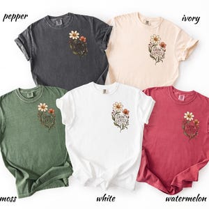 May include: Five short-sleeved crew neck t-shirts in pepper, ivory, moss, white, and watermelon colours. Each shirt features a small floral graphic on the left chest with the text "GROW GENTLY" printed in a script font amidst brown and white flowers.