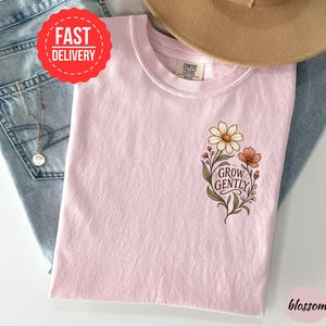 May include: A light pink t-shirt with a Comfort Colors label at the neck. The shirt features a graphic on the chest depicting a cluster of flowers in shades of cream, orange, and green, with the text "GROW GENTLY" written in a dark script font.