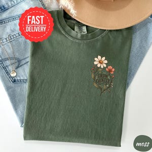 May include: A moss green crew neck t-shirt featuring a small floral graphic on the left chest. The design includes white and pink flowers with green leaves and the text "GROW GENTLY" in a dark script font. The shirt has a visible brand tag.