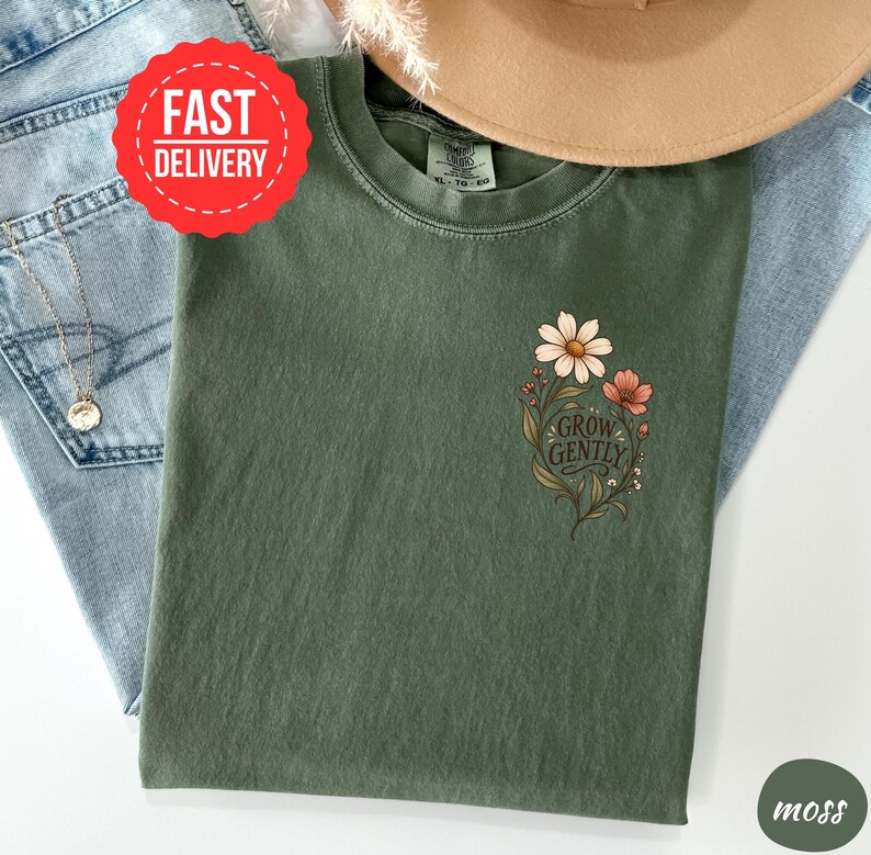 May include: A moss green crew neck t-shirt featuring a small floral graphic on the left chest. The design includes white and pink flowers with green leaves and the text "GROW GENTLY" in a dark script font. The shirt has a visible brand tag.