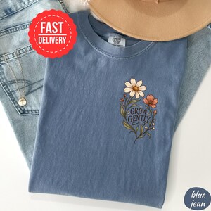May include: A blue short-sleeved t-shirt featuring a graphic of two flowers and green leaves with the text "GROW GENTLY" printed in a dark script font. The shirt has a crew neckline and a label that reads "COMFORT COLORS" at the inner collar.