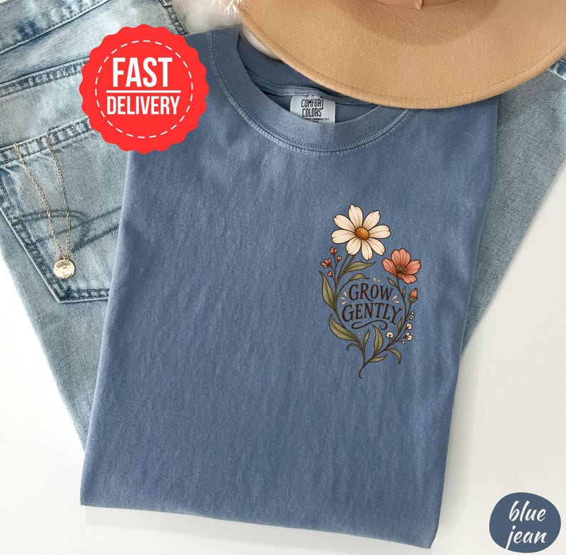 May include: A blue short-sleeved t-shirt featuring a graphic of two flowers and green leaves with the text "GROW GENTLY" printed in a dark script font. The shirt has a crew neckline and a label that reads "COMFORT COLORS" at the inner collar.