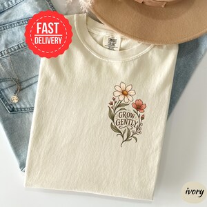 May include: An ivory t-shirt featuring a graphic of flowers and the text "GROW GENTLY" in a script font. The shirt has a crew neckline and a Comfort Colors brand label at the inner collar. The design includes green leaves and stems with pink and white petals.