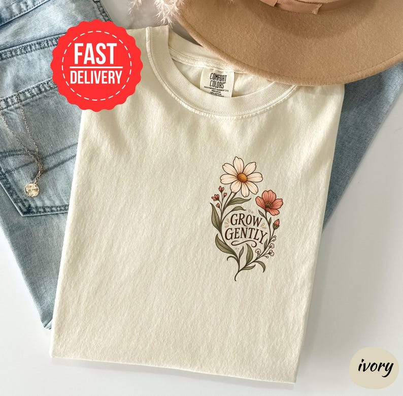 May include: An ivory t-shirt featuring a graphic of flowers and the text "GROW GENTLY" in a script font. The shirt has a crew neckline and a Comfort Colors brand label at the inner collar. The design includes green leaves and stems with pink and white petals.