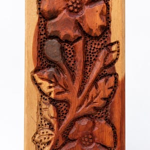 May include: A rectangular wooden plaque featuring a hand-carved floral design. The relief carving displays two prominent flowers and leaves set against a dark, stippled background. The wood exhibits natural grain patterns with light and dark brown tones.