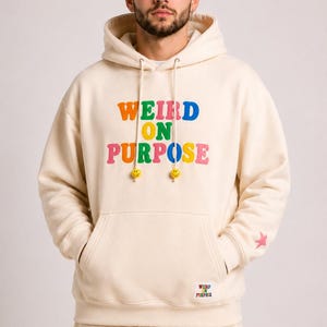 May include: A cream-colored hooded sweatshirt featuring the text WEIRD ON PURPOSE in multicolored letters on the chest. The hoodie includes a front kangaroo pocket, drawstring cords with yellow smiley face beads, and a small pink star on the sleeve.