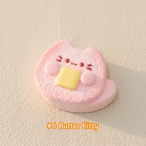 May include: A light pink, textured cat-shaped figurine featuring a small, rectangular yellow butter pat on its chest. The cat has a simple face with two dark brown eyes, a small mouth, and three whiskers on each side. The text #6 Butter Kitty is below.