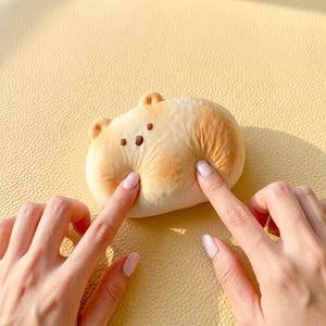 May include: A soft, squishy, beige-colored toy shaped like a capybara with small, rounded ears and three dark brown dots representing eyes and a nose. The surface of the toy is textured and shows visible indentations from being pressed.