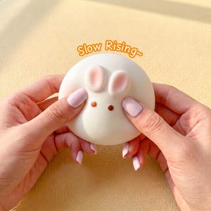 May include: A round, white, squishy toy shaped like a rabbit with two small pink ears and two brown dot eyes. The text "Slow Rising~" is printed in orange above the toy. The object has a smooth, matte texture and is held between two hands.