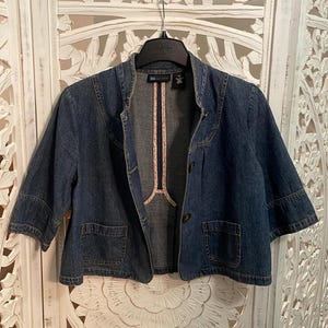 May include: A dark blue denim jacket with three-quarter length sleeves, a mandarin collar, and two front patch pockets. The jacket features a button-front closure and a light-colored floral trim along the inner placket and neckline seams.