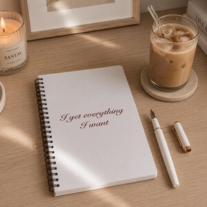 Può includere: Un quaderno a spirale bianco con il testo I get everything I want stampato in corsivo marrone scuro sulla copertina. Il quaderno è posizionato accanto a una penna stilografica bianca con dettagli dorati e al suo cappuccio abbinato.