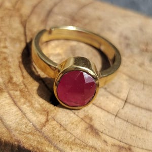 Könnte beinhalten: Ein goldfarbener Ring mit einem runden, facettierten, dunkelroten Edelstein in einer Zargenfassung. Die Ringschiene ist glatt und weist oben eine kleine Öffnung auf, was auf ein verstellbares Design hindeutet. Das Metall hat eine polierte Oberfläche.