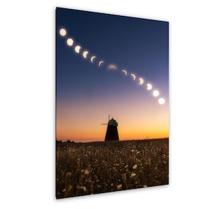 Puede incluir: Una impresión de arte mural vertical que muestra una secuencia de eclipse solar formando un arco en un cielo crepuscular azul oscuro y naranja sobre la silueta de un molino de viento tradicional en un campo de flores silvestres.