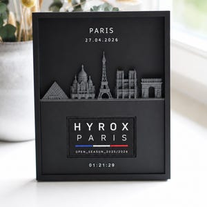 Könnte beinhalten: Ein schwarzer, rechteckiger Schattenkasten-Rahmen mit einer grauen 3D-Skyline von Pariser Wahrzeichen, darunter der Eiffelturm. Der Text lautet PARIS 27.04.2026, HYROX PARIS, OPEN_SEASON_2025/2026 und 01:21:29, mit einem blau-weiß-roten Streifen.