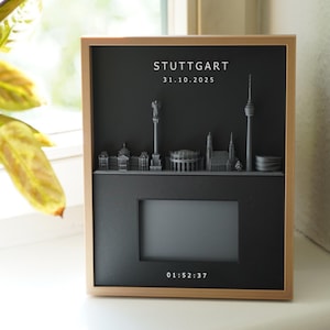 Könnte beinhalten: Ein rechteckiges Wandbild mit hellem Holzrahmen und schwarzem Hintergrund. Es zeigt eine graue 3D-Skyline von Stuttgart, den Text STUTTGART 31.10.2025, einen zentralen rechteckigen Ausschnitt für ein Foto und die digitale Uhrzeit 01:52:37 unten.