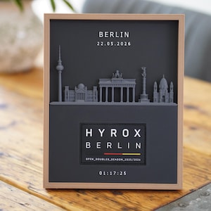 Könnte beinhalten: Ein gerahmtes Wandbild mit einer grauen 3D-Silhouette der Berliner Skyline. Der Text lautet BERLIN, 22.05.2026, HYROX BERLIN, OPEN_DOUBLES_SEASON_2025/2026 und 01:17:25. Unter dem Text befindet sich eine kleine Grafik der deutschen Flagge.