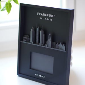 Könnte beinhalten: Ein schwarzer Schattenfugenrahmen mit einer grauen 3D-Skyline von Frankfurt. Der Text FRANKFURT und das Datum 14.12.2025 stehen in Weiß über der Skyline, während die Uhrzeit 02:21:52 unten unter einem rechteckigen Ausschnitt in Weiß zu sehen ist.