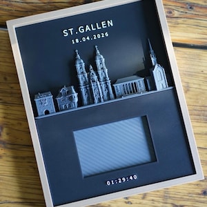 Könnte beinhalten: Ein rechteckiger Schattenfugenrahmen in hellem Holz mit schwarzem Hintergrund. Er zeigt eine graue 3D-Skyline von St. Gallen, den Text ST.GALLEN 18.04.2026, einen zentralen rechteckigen Ausschnitt für ein Foto und die Uhrzeit 01:29:40 darunter.