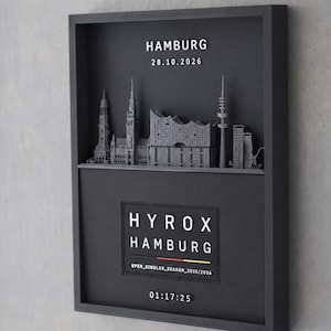 Könnte beinhalten: Ein schwarzes Wandbild mit einer grauen 3D-Skyline von Hamburg, dem Text HAMBURG 28.10.2026 und einer schwarzen Plakette mit der Aufschrift HYROX HAMBURG, OPEN_SINGLES_SEASON_2025/2026, einer deutschen Flagge und der Zeitangabe 01:17:25.