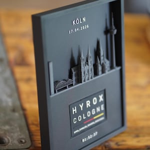 Könnte beinhalten: Eine mattschwarze, 3D-gedruckte Wandtafel mit einem Relief der Kölner Skyline, dem Text KÖLN, 17.04.2026 sowie einem rechteckigen Einsatz mit dem Text HYROX COLOGNE, einer kleinen deutschen Flagge und der Zeitangabe 01:52:37 am unteren Rand.