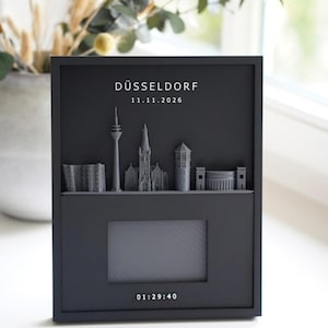 Könnte beinhalten: Ein schwarzer rechteckiger Rahmen mit einer grauen 3D-Skyline von Düsseldorf, dem Text DÜSSELDORF, dem Datum 11.11.2026, einem zentralen rechteckigen Ausschnitt für ein Foto und der Uhrzeit 01:29:40, die unten auf dem Rahmen weiß aufgedruckt ist.