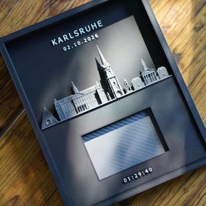 Könnte beinhalten: Ein schwarzer Wandbilderrahmen mit einer silbernen 3D-Skyline von Karlsruhe. Der Text lautet KARLSRUHE, 02.10.2026 und 01:29:40. Ein rechteckiger Ausschnitt in der Mitte zeigt ein Karbonfasermuster und bildet eine moderne Darstellung der Architektur.