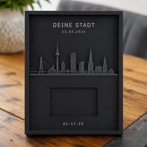 Könnte beinhalten: Ein schwarzer rechteckiger Wandrahmen mit einem 3D-Relief einer Stadtsilhouette. Der Text DEINE STADT und 22.05.2026 stehen oben, mit einem rechteckigen Ausschnitt in der Mitte und den in Weiß gedruckten Zahlen 01:17:25 am unteren Rahmenrand.