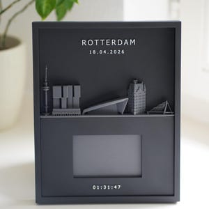 Könnte beinhalten: Ein schwarzer rechteckiger Rahmen mit einer grauen 3D-Skyline von Rotterdam. Der Text "ROTTERDAM" und "18.04.2026" steht oben, während "01:31:47" unten aufgedruckt ist. Ein zentraler rechteckiger Ausschnitt befindet sich zwischen Skyline und Zeit.