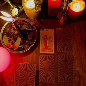 Pode incluir: Uma disposi&ccedil;&atilde;o de cartas de tarot com a carta Oito de Espadas virada para cima, rodeada por v&aacute;rias cartas viradas para baixo com um padr&atilde;o de raios, uma ta&ccedil;a de metal com ervas secas e uma vela acesa, e v&aacute;rias velas de pilar acesas.