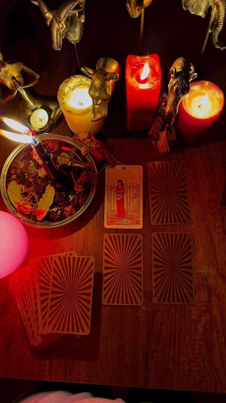 Pode incluir: Uma disposi&ccedil;&atilde;o de cartas de tarot com a carta Oito de Espadas virada para cima, rodeada por v&aacute;rias cartas viradas para baixo com um padr&atilde;o de raios, uma ta&ccedil;a de metal com ervas secas e uma vela acesa, e v&aacute;rias velas de pilar acesas.