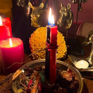 Pode incluir: Uma ta&ccedil;a de metal prateado cheia de ervas secas, folhas e um pau de canela, contendo duas velas verticais, uma preta e uma vermelha, atadas com um fio vermelho. Uma vela pilar vermelha grande est&aacute; &agrave; esquerda, com figuras de animais atr&aacute;s.