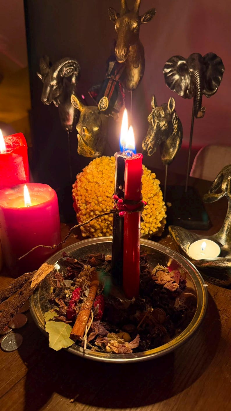 Pode incluir: Uma ta&ccedil;a de metal prateado cheia de ervas secas, folhas e um pau de canela, contendo duas velas verticais, uma preta e uma vermelha, atadas com um fio vermelho. Uma vela pilar vermelha grande est&aacute; &agrave; esquerda, com figuras de animais atr&aacute;s.